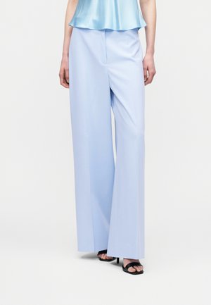 Γυναίκα φοράει ανοιχτό μπλε παντελόνια με φαρδύ πόδι, σατινέ ανοιχτό μπλε τοπ χωρίς μανίκια και μαύρα ψηλοτάκουνα σανδάλια με ανοιχτές μύτες, στέκεται μπροστά σε λευκό φόντο.