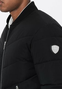 Svart uppblåsbara jacka med dragkedja framtill, ribbad krage och en silverfärgad logotyp patch på vänster ärm. Har en kviltad struktur och djärv sömnad.