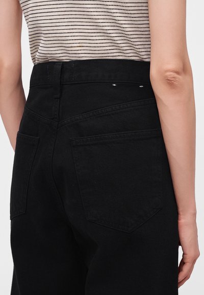 Femme portant un jean noir taille haute et un top sans manches beige et noir rayé, vue de dos et de la taille vers le bas.