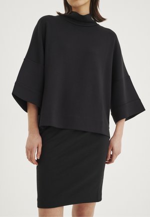 Černý oversized top s vysokým límcem a širokými rukávy, který je spárován s upnutou černou sukní, obě vyrobené z hladkého materiálu.