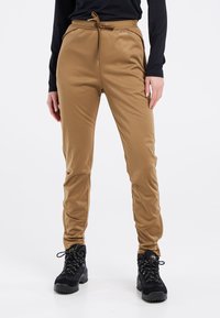 Tan, slim-fit sportieve broek met een elastische tailleband, zijzakken en een gladde textuur, gecombineerd met zwarte wandelschoenen.