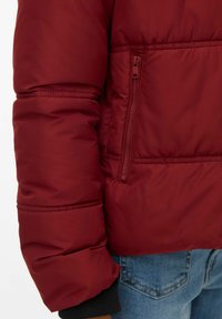 Burgunderfarget polstret jakke med sidelomme med glidelås, ribbestrikkede mansjetter og glatt stofftekstur. Figursydd passform med quiltede detaljer.