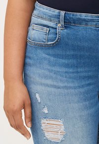 Close-up of a hand resting on the thigh of distressed denim jeans, featuring a rip and faded blue fabric.