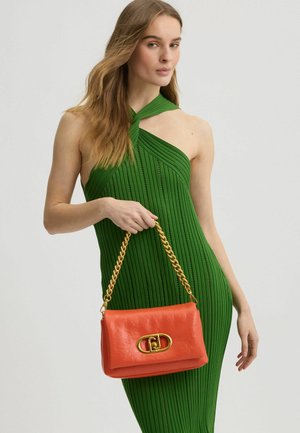 Helle orange Lederhandtasche mit einer goldenen Kettenschlaufe, ausgestattet mit einem markanten ovalen Verschluss. In Szene gesetzt vor einem Modell, das ein grünes geripptes Kleid trägt.