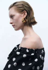 Cercei circulare de aur, bluză neagră cu buline, cu umerii la vedere, păr neted, fundal neutru, vedere din profil a unei persoane.