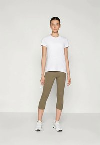 Biely tričko s krátkym rukávom v kombinácii s olivovozelenými legínami do polovice lýtok a bielymi športovými teniskami. Neutrálny pozadie zdôrazňuje oblečenie.