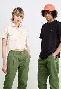 Cropped beige button-up shirt with two pockets, paired with green pants. Black t-shirt with a small logo and orange cap on the right.