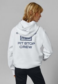 Blondín v bielej mikine s kapucňou, na zadnej strane s textom "RBR PIT STOP CREW" a čiernymi nohavicami, stojaci proti šedému pozadiu.