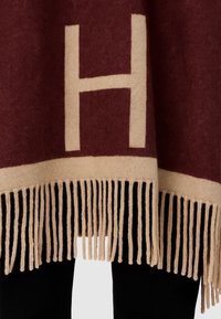 Coperta color bordeaux con motivo "H" crema, caratterizzata da un bordo frangiato. Texture morbida, piegata parzialmente su un tessuto nero per creare contrasto.