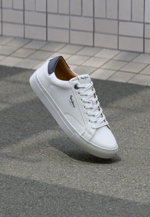 Sneaker bianco in pelle con una finitura liscia, tomaia con un tallone grigio a contrasto e suola in gomma testurizzata. Presenta lacci piatti e un dettaglio del logo sul lato.
