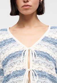 Γυναίκα που φοράει πλεκτό πουλόβερ μπλε και λευκό με δύο κορδόνια μπροστά, αποκαλύπτοντας το λαιμό της, τις κλείδες και μέρος του προσώπου και των μαλλιών της.