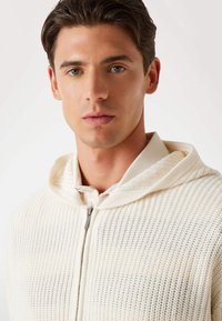 Leichte, cremefarbene, gestrickte Kapuzenjacke mit Reißverschluss, gerippten Bündchen und einer strukturierten Stoffoberfläche. Hat eine lockere Passform und eine Kapuze.