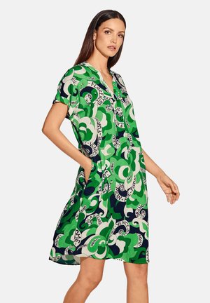 Frau trägt ein kurzärmeliges, knielanges Kleid mit grünem, marineblauem und weißem Muster mit Städtenamen in wirbelndem Design, steht mit einer Hand in der Tasche.