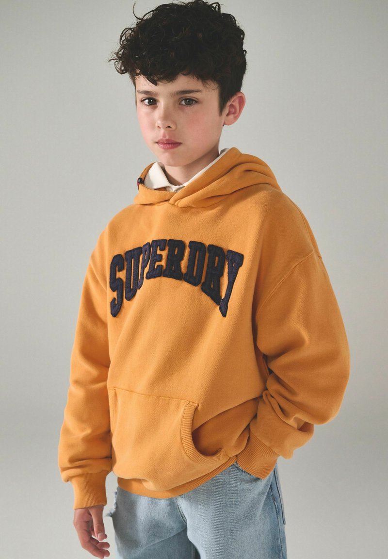 Niño con cabello rizado y oscuro que lleva una sudadera amarillo mostaza de Superdry y jeans azul claro, apoyado contra un fondo gris sencillo.