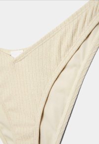 Gros plan du bas de bikini en tissu côtelé crème montrant la texture et les coutures sur les côtés intérieur et extérieur.