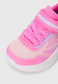 Chaussure de sport rose avec une tige en maille, des lacets élastiques et une sangle Velcro. La semelle blanche présente des accents texturés et un logo sur la sangle.