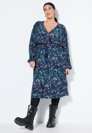 Kvinde iført en blå blomster-midi-kjole med lange ærmer, sorte leggings og chunky sorte støvler til midt på læggen, stående mod en hvid baggrund.