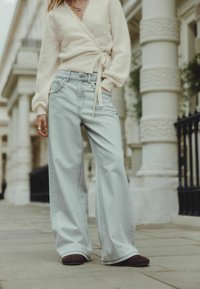 Világoskrém színű, derekán megkötővel ellátott kardigán, ami egy világoskék, bő szabású farmerral és sötétbarna cipőkkel van párosítva.