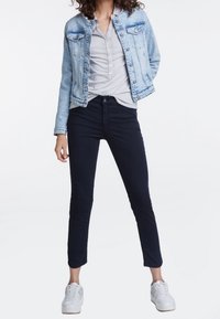 Persona vestida con una chaqueta de mezclilla azul claro, camisa gris de botones, pantalones negros ajustados y zapatillas blancas, de pie contra un fondo blanco liso.