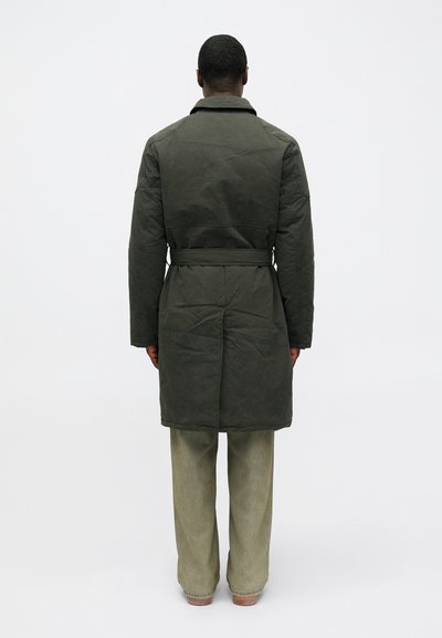 Palton careptat de culoare verde închis, cu curea, mâneci lungi și guler. Are o textură netedă. Se poartă peste pantaloni largi de culoare verde deschis.