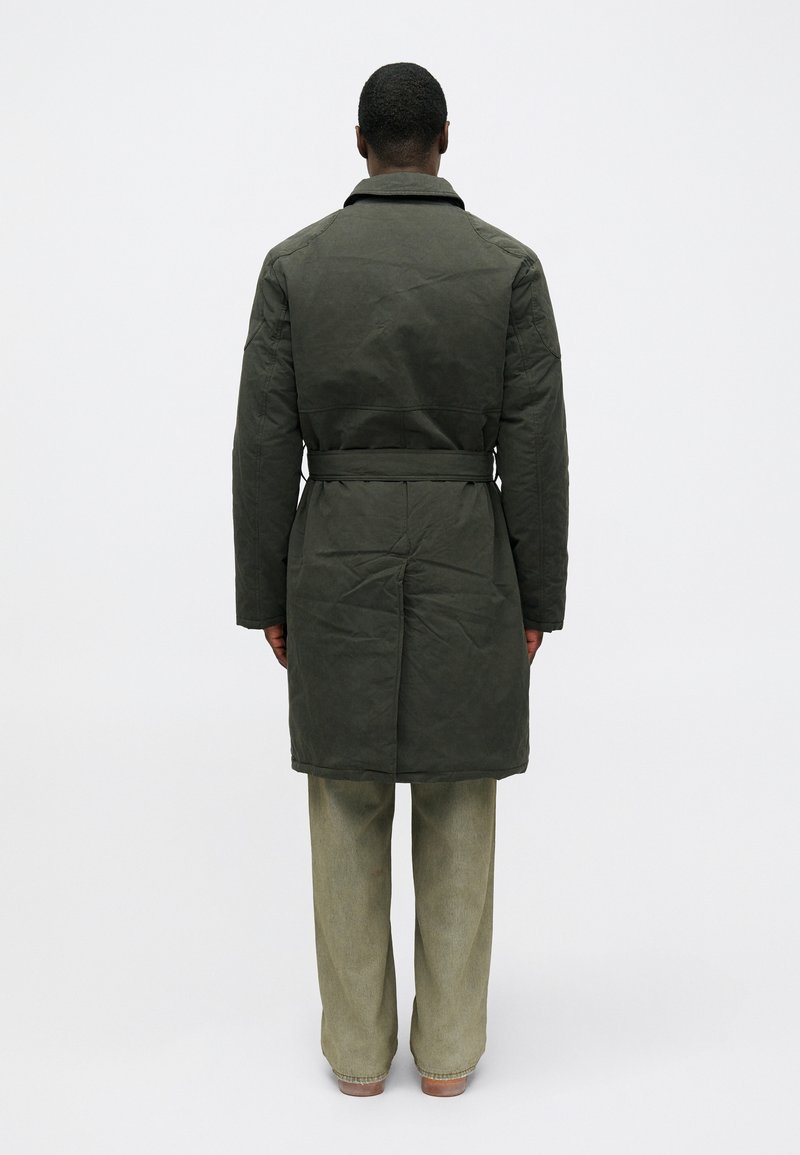 Donkergroen gewatteerde jas met een riem, lange mouwen en een kraag. Heeft een gladde textuur. Gede gedragen over lichtgroene wijde broek.