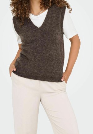 Personne portant un t-shirt blanc à manches courtes sous un gilet sans manches en tricot marron foncé et un pantalon beige clair, les mains dans les poches, sur fond blanc.