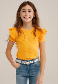 Dzeltena ribota īsām piedurknēm krekls ar frēzēm, apvienots ar zilām džinsām un teksturētu sudraba jostu ar izgriezumu dizainiem.