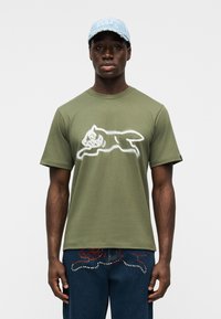 Λαδί βαμβακερό T-shirt με λευκό σχέδιο ενός άλματος ζώου. Συνδυάζεται με μπλε τζιν τζιν με διακοσμητικές λεπτομέρειες.