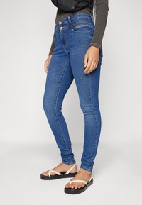 Közepes kék farmer skinny farmert visel, magas derékkal, kontrasztvarrással és két gombos záródással. Fehér strandpapuccsal viseli.