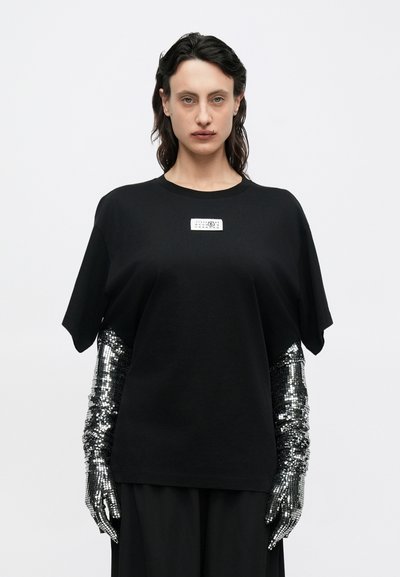 Μαύρο φαρδύ T-shirt με κοντά μανίκια, που διαθέτει ένα ορθογώνιο ετικέτα στο εμπρός μέρος. Συνοδεύεται από ασημένια καλύμματα χεριών με παγιέτες.