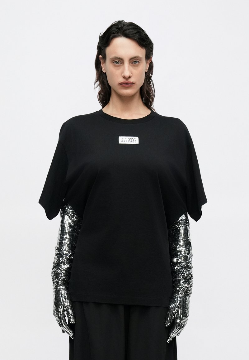 Schwarzes, übergroßes T-Shirt mit kurzen Ärmeln und einem rechteckigen Etikett auf der Vorderseite. Dazu silberne Armstulpen mit Pailletten.