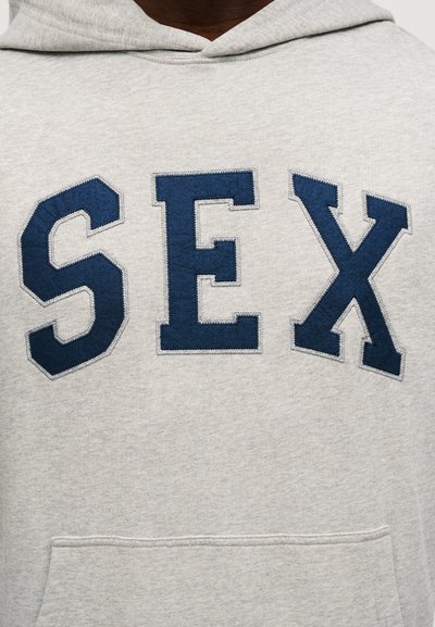 Pilkas džemperis su gobtuvu, priekyje esančiais dideliais mėlynais siuvinėtais raidėmis, kurios sudaro žodį „SEX“, ir priekinė kišenė.