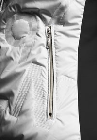 Witte donsjas met een ritszak, een gestructureerd patroon en een glanzende zilveren rits op een contrasterende zwarte achtergrond.