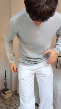 Άνδρας που φοράει ανοιχτό γκρι μακρυμάνικο πουκάμισο και λευκό παντελόνι, προσαρμόζοντας τα ρούχα του ενώ στέκεται σε ένα δωμάτιο με κρεβάτι και βιβλία κοντά.