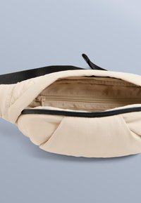 Beige midjeväska i nylon med ett vikbart design, svart blixtlås och en innerficka. Har en svart justerbar rem för säker användning.