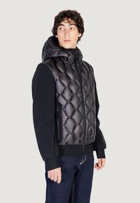 Giacca nera imbottita con texture trapuntata, dotata di colletto alto e cappuccio, polsini elasticizzati e tasca laterale con zip.