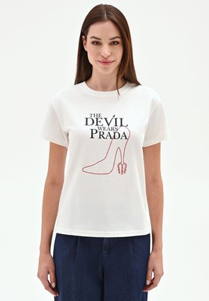 Donna con capelli castani lunghi che indossa una maglietta bianca con la scritta "The Devil Wears Prada" e una grafica di tacchi rossi, abbinata a pantaloni blu scuro.