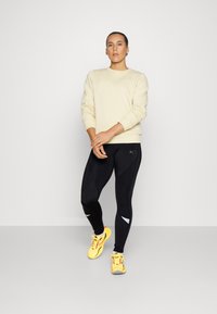 Felpa beige chiaro, leggings neri con dettagli testurizzati e scarpe da corsa gialle brillanti con lacci rossi; posa in piedi.