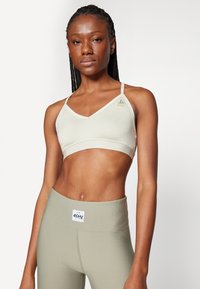 Sujetador deportivo verde claro de tejido suave y elástico, con escote en V y tirantes finos. Presenta un logotipo de marca en el lado izquierdo. Combinado con leggings de oliva de alta cintura y con canalé.