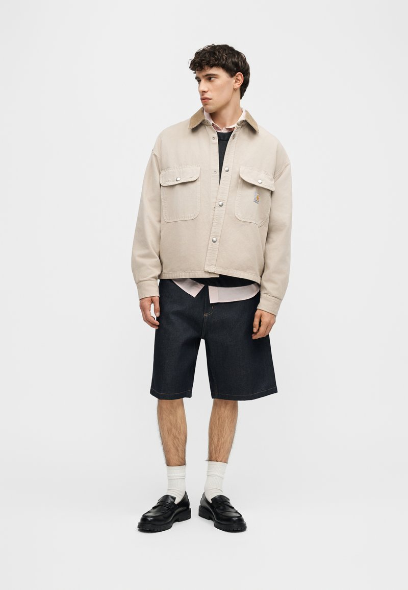 Joven con chaqueta beige sobre suéter negro y camisa clara, pantalones cortos oscuros hasta la rodilla, calcetines blancos y mocasines negros, de pie sobre fondo blanco.