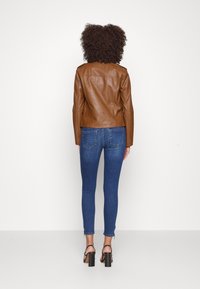 Veste en cuir marron de style ajusté, avec deux épaulettes, associée à un jean skinny bleu. Chaussures noires à talons carrés portées.