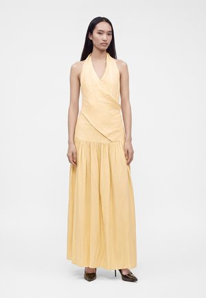 Femme aux longs cheveux foncés portant une robe longue sans manches jaune pâle à col halter et des chaussures à talons hauts noires sur un fond blanc uni.