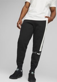 Svarta sweatpants i bomull med ett vitt sidostrick, med en logotyp nära fållen. De har en figurnära passform och ribbade muddkanter.
