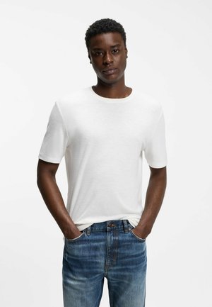 Giovane uomo con capelli corti e ricci, indossa una t-shirt bianca a maniche corte semplice e jeans blu con le mani nelle tasche, sfondo neutro.