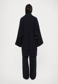Námořnický vlněný kabát s širokým siluetou, oversized rukávy a vysokým límcem, spárovaný s pruhovanými kalhotami. Minimalistický design, hladká textura.