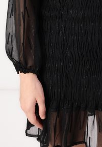 Svart klänning med genomskinligt tyg som har en texturerad smockad design och elastiska puffärmar. Noterbara reflekterande detaljer överallt.