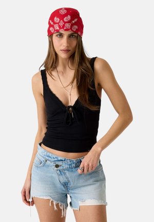 Frau mit rotem, gemustertem Bandana, schwarzem ärmellosem Top mit vorderer Schleife und hellblauen, verwaschenen Jeansshorts, stehend vor weißem Hintergrund.