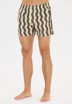 Barbenet person iført beige og sorte bølgede stribede badeshorts med hvid snor, stående mod en ensfarvet hvid baggrund.