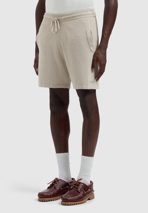 Man draagt beige textuurige korte broek met aantrekkoord, witte sokken en bruine leren bootschoenen, staand tegen een effen achtergrond.