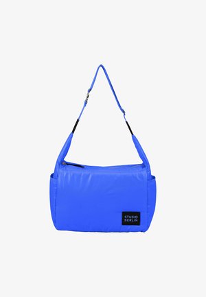 Sac à main rembourré bleu avec une bandoulière réglable, fermeture éclair en haut et une poche latérale. Il comporte un écusson noir avec l'inscription "STUDIO BERLIN."